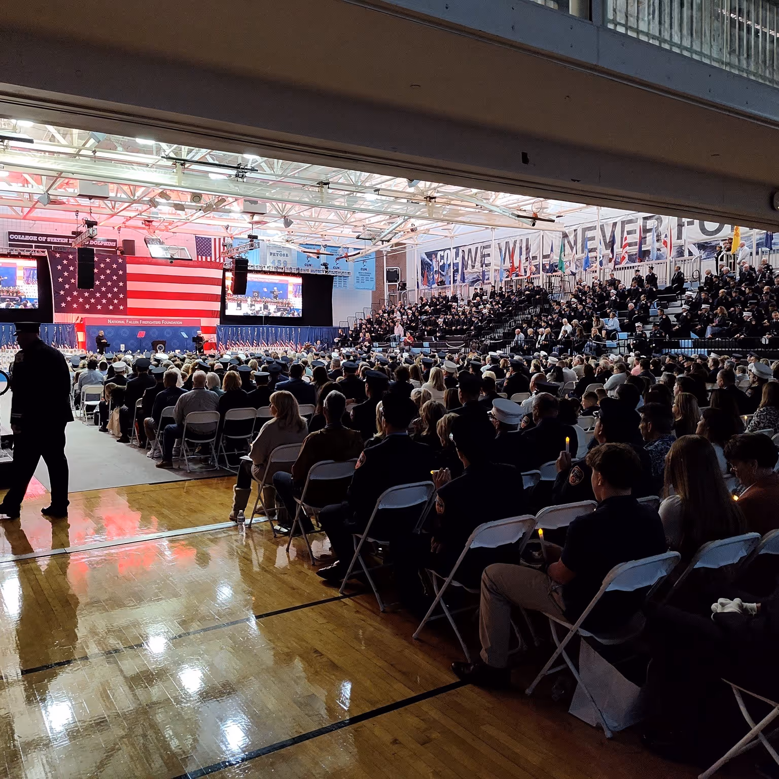 Large indoor gathering with an American flag backdrop and seated attendees, many in uniform.