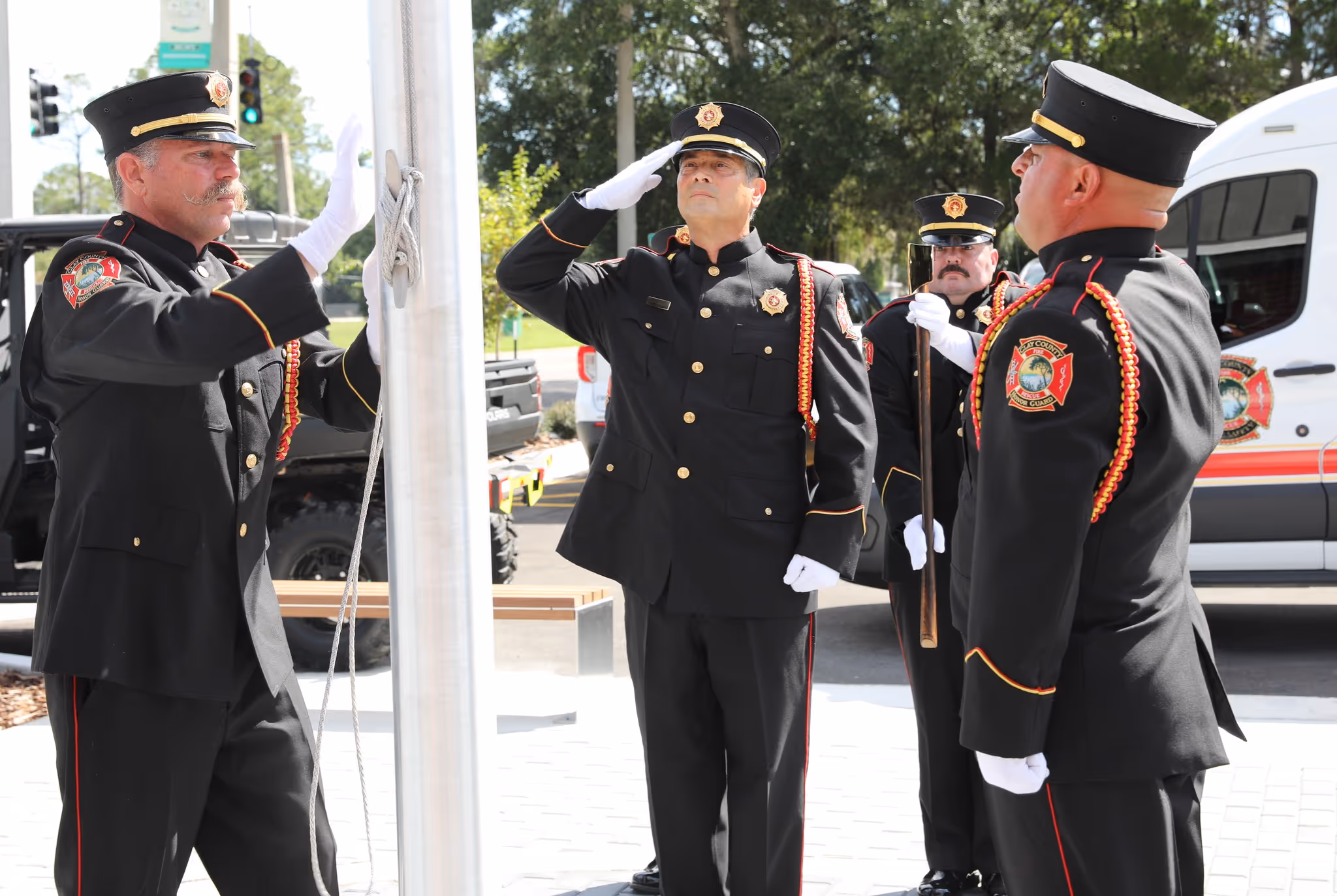 Four uniformed honor guard members in black dress coats performing a flag ceremony outdoors.