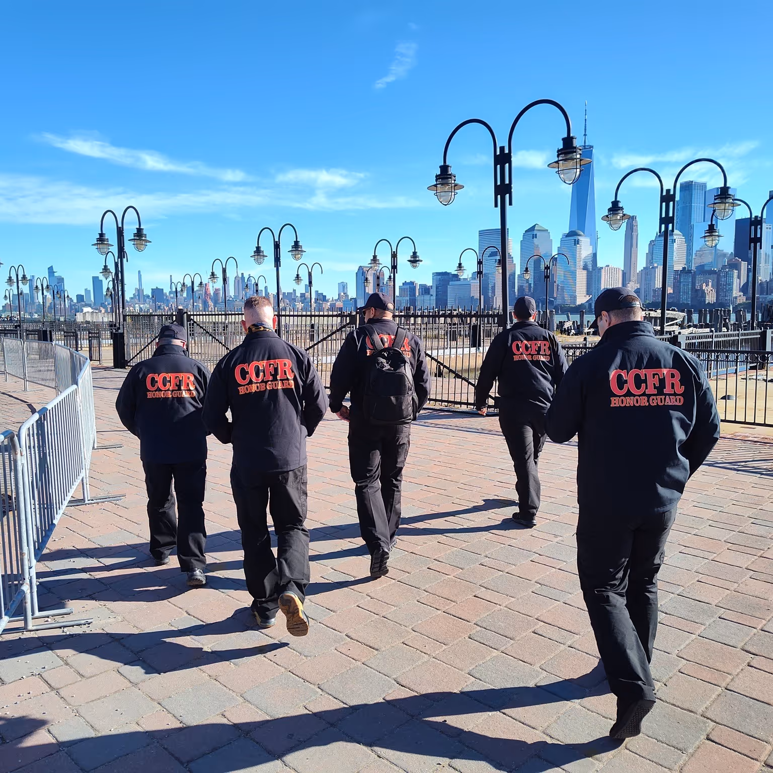 Five people in CCFR Honor Guard jackets walking on paved path with city skyline and lampposts in background.