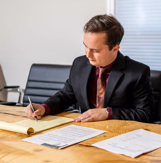 A man in a black suit and maroon shirt with a patterned tie is seated at a wooden table, writing on a notepad with a pen, focused on his work.