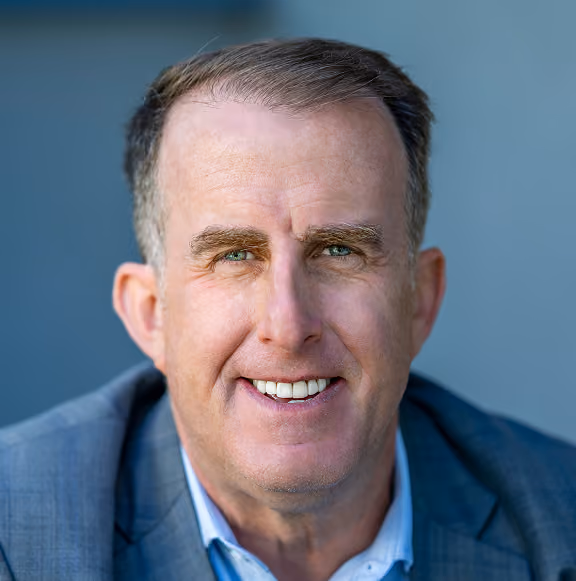 Close-up portrait of a middle-aged man with short brown hair, green eyes, wearing a grey suit jacket and light blue shirt, smiling against a blurred blue background.