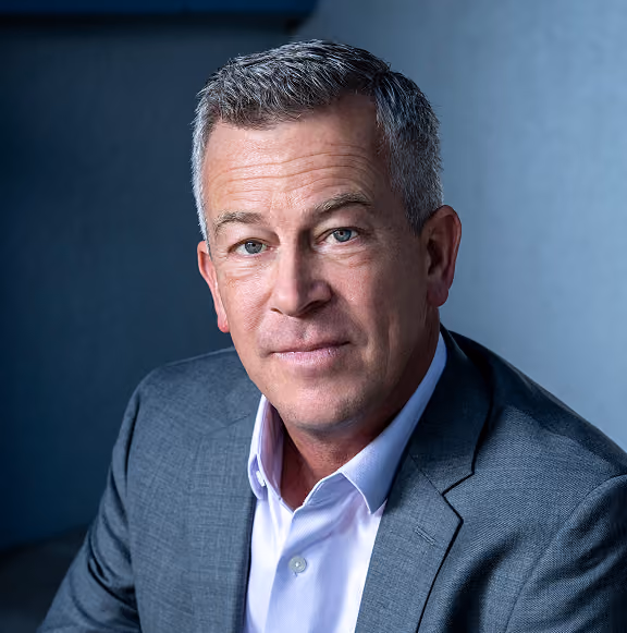 Portrait of a middle-aged man with short salt-and-pepper hair wearing a gray suit jacket and white shirt, looking directly at the camera with a slight smile, against a blurred blue background.