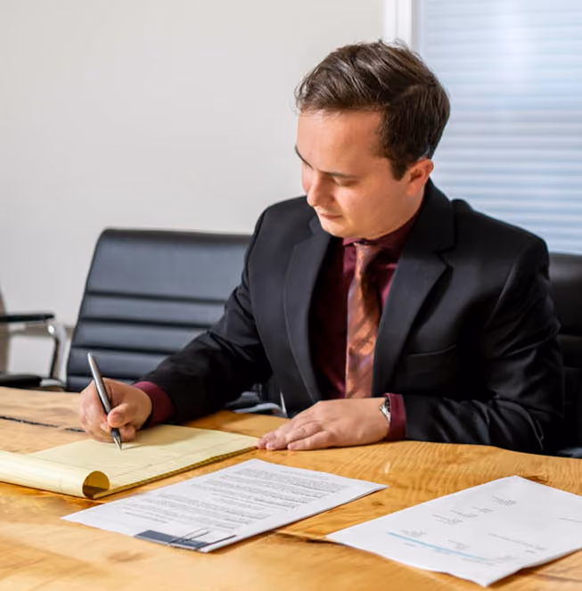 A man in a black suit and maroon shirt with a patterned tie is seated at a wooden table, writing on a notepad with a pen, focused on his work.