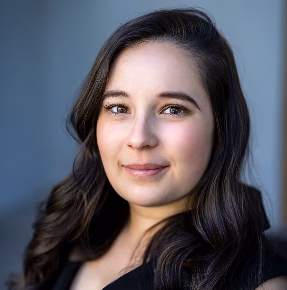 Portrait of a young woman with long dark wavy hair, light skin, and hazel eyes, wearing a black top, smiling softly against a blurred gray background.