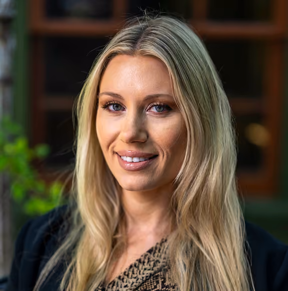 Portrait of a woman with long blonde hair smiling gently, wearing a patterned top and a black jacket, with a blurred natural background.