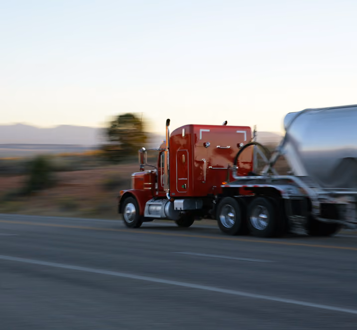Red semi-truck hauling a shiny silver tanker trailer driving quickly on a highway with blurred background.