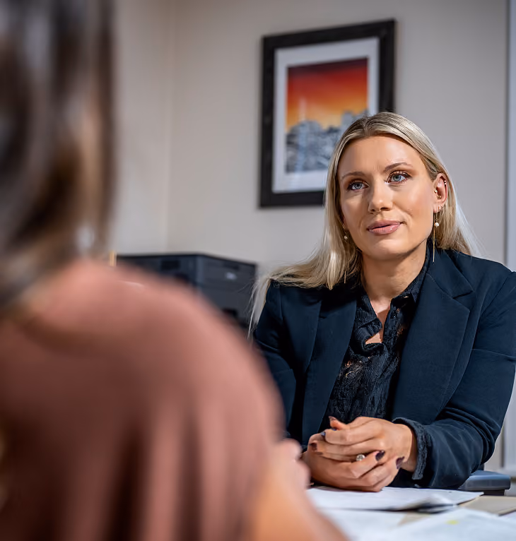 A professional blonde woman in a dark blazer attentively listens to another person across a desk in an office setting.