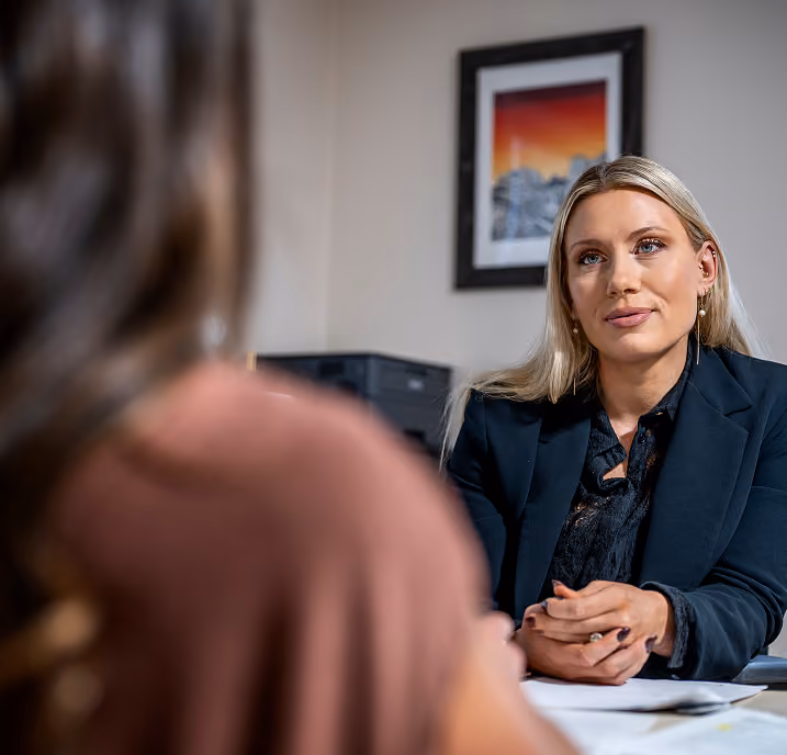 A professional woman with blonde hair, wearing a dark blazer, attentively listening during a conversation or meeting in an office setting, with the blurred back of another person in the foreground and a framed picture on the wall behind her.