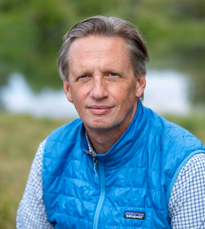 Middle-aged man with light brown hair wearing a blue Patagonia quilted vest over a checkered shirt, sitting outdoors with a blurred green background.