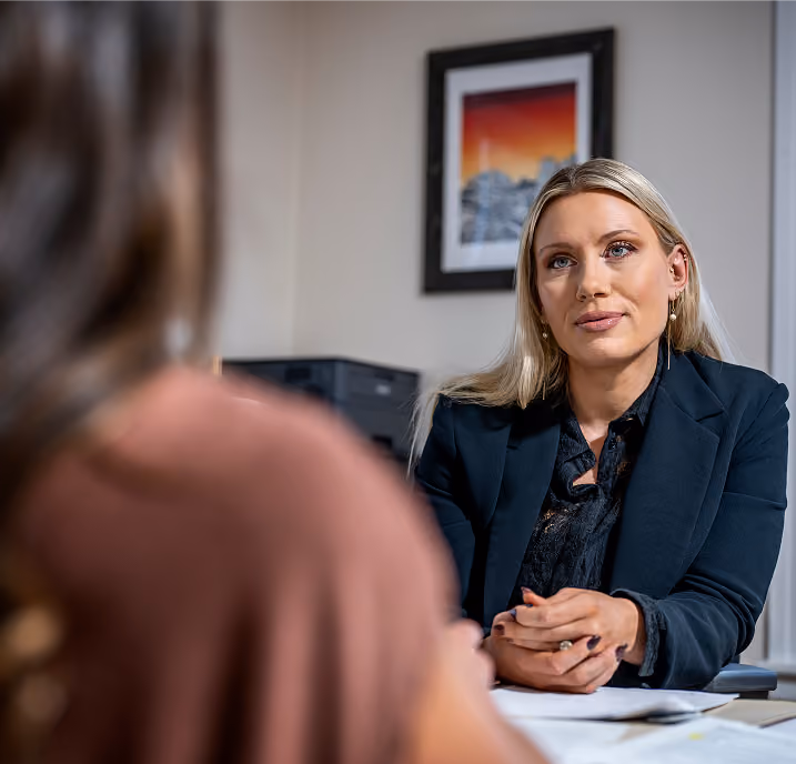 A blonde woman in a dark blazer attentively listens and engages in conversation with another person in an office setting, with a framed picture on the wall behind her.