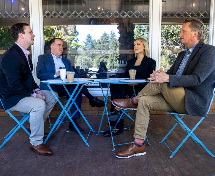 Four business professionals, two men on the left and a woman and man on the right, sitting on blue metal chairs around two blue metal tables outside a building, each with iced coffee cups, engaged in conversation.
