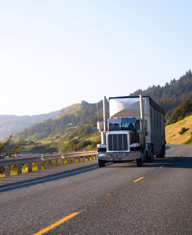A large semi-truck with a white trailer driving on a two-lane highway surrounded by green hills and trees under a clear sky.