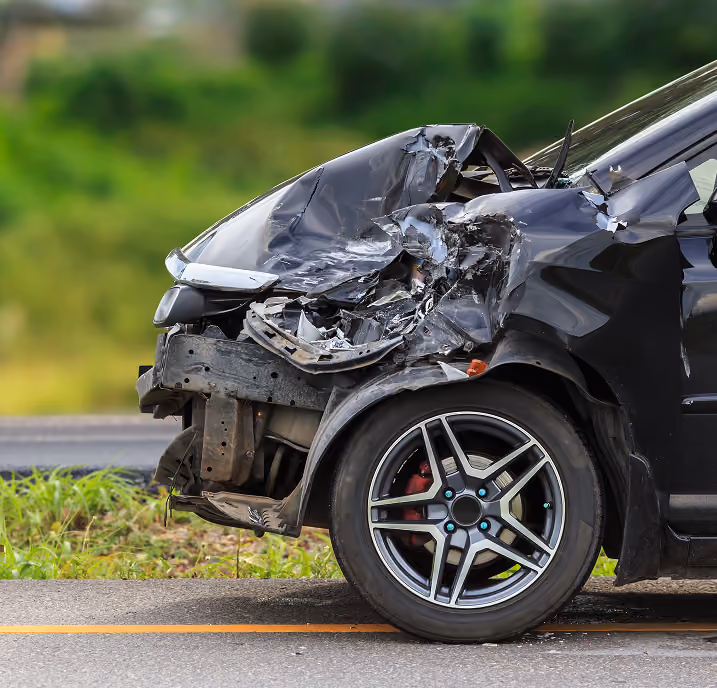Front-left side of a black car severely damaged with a crumpled fender, broken headlight, and exposed internal parts, parked by a roadside with greenery in the background.