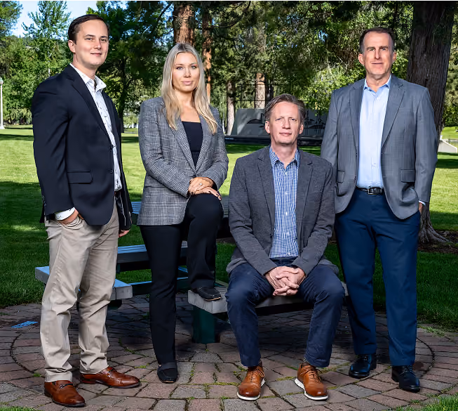 Group of four professionally dressed adults posing outdoors on a circular stone patio with green grass and trees in the background; two men stand on either side, a man sits on a picnic bench, and a woman rests one foot on the bench.