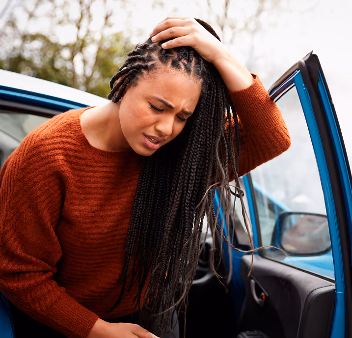 Woman with long braided hair wearing a rust-colored sweater leans forward next to an open blue car door, holding her head with a pained expression.