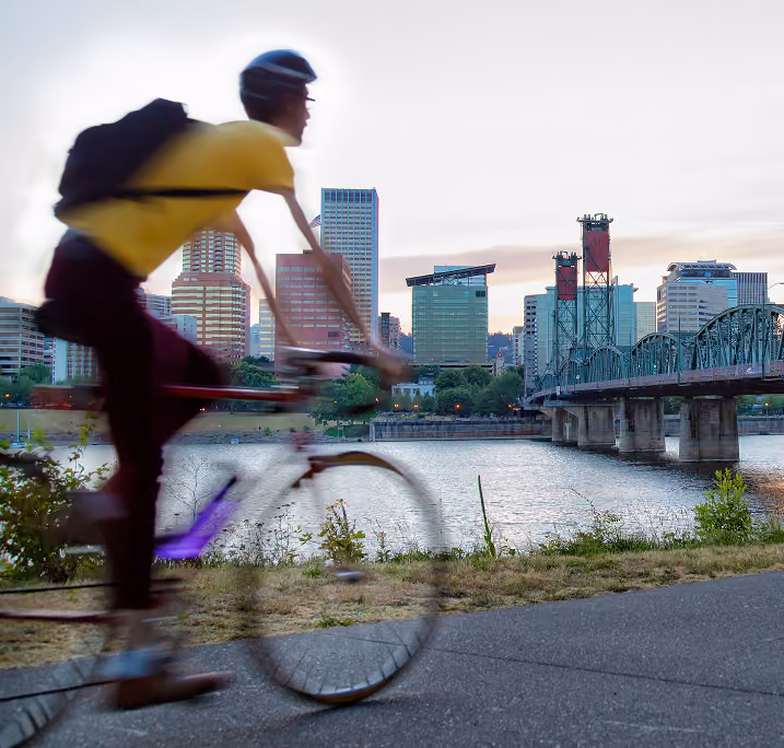 Person wearing a yellow shirt and helmet rides a bicycle on a path along a river with a city skyline and a green steel bridge in the background at dusk.
