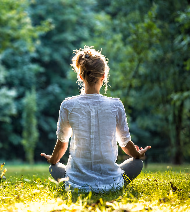 Person sitting cross-legged on grass outdoors in a meditative pose facing away, surrounded by green trees.
