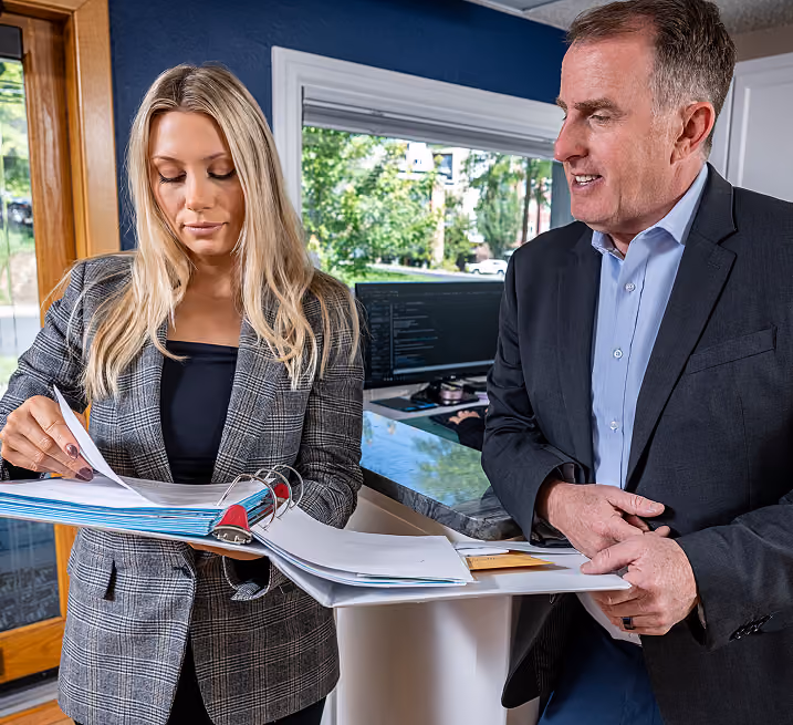 A man in a dark suit and blue shirt is talking to a woman in a gray plaid blazer who is reviewing papers in a binder, standing in an office with a window showing green trees outside and a computer monitor in the background.