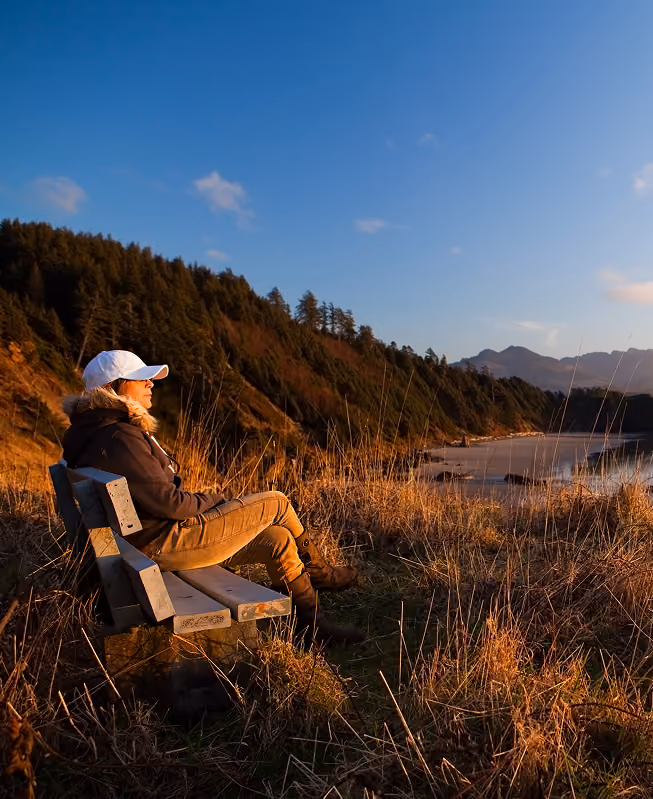 Person wearing a white cap and brown jacket sits on a wooden bench facing a lake at sunset, with forested hills and mountains in the background under a clear blue sky.