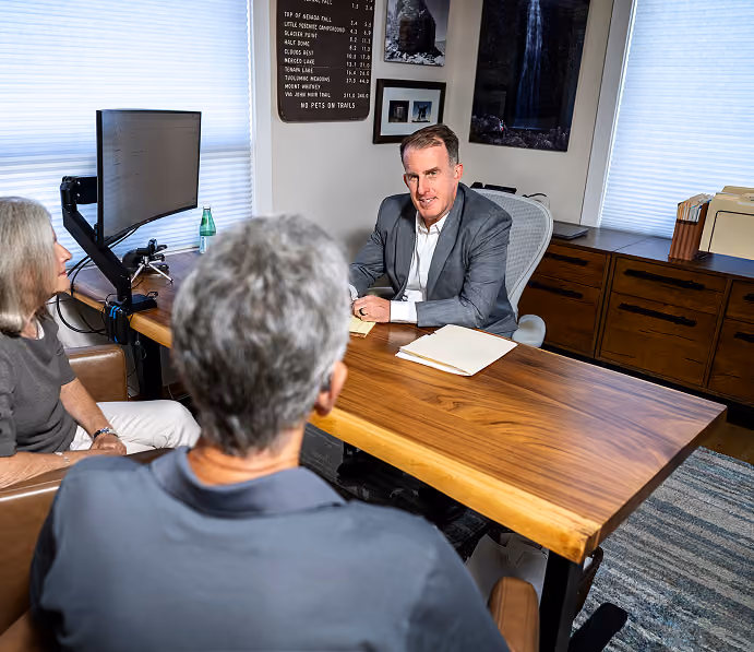 A man in a gray suit sitting at a wooden desk talking to an older woman and man seated across from him in an office setting with a computer monitor on the desk.