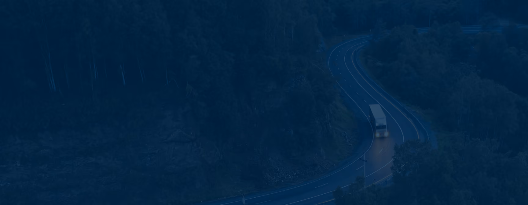 A lone truck driving on a winding road surrounded by dense, dark forested hills at dusk or night.
