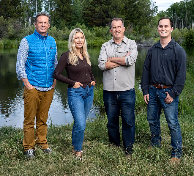Four adults standing outdoors on grass near a calm pond with trees in the background. From left to right: a man in a blue vest and brown pants, a blonde woman in a black top and blue jeans, a man in a light gray shirt and dark jeans with arms crossed, and a man in a black shirt and blue jeans with a brown belt. All are smiling and facing forward.