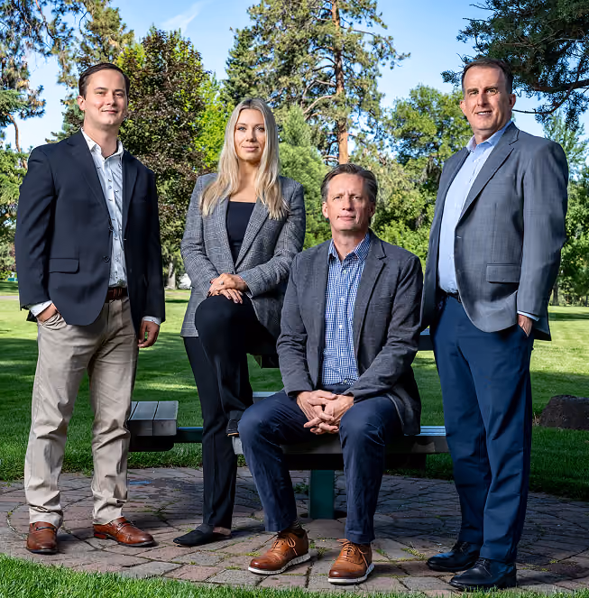 Group of four professionally dressed adults posing outdoors on a circular stone patio with green grass and trees in the background; two men stand on either side, a man sits on a picnic bench, and a woman rests one foot on the bench.