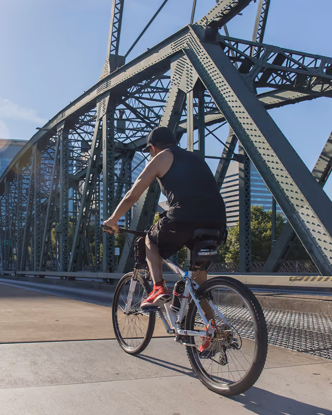 Person wearing black tank top, shorts, and red shoes riding a bicycle on a steel truss bridge under clear blue sky.