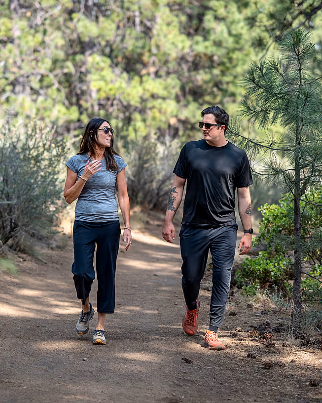 A man and a woman wearing sunglasses walking and talking on a wooded trail surrounded by trees and bushes.