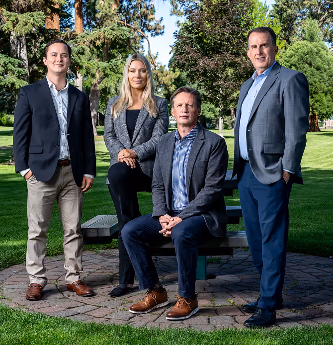 Group of four professionally dressed adults posing outdoors on a circular stone patio with green grass and trees in the background; two men stand on either side, a man sits on a picnic bench, and a woman rests one foot on the bench.