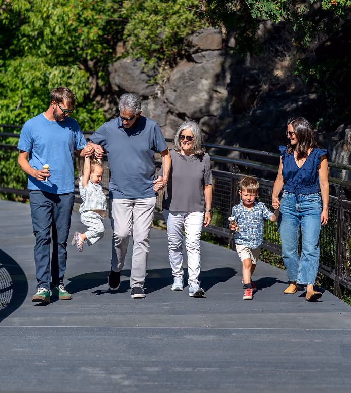 Five people walking outdoors on a sunny day on a paved path with greenery and rocks in the background; an older man and woman hold hands with two young children, the man swinging one child while a young woman holds the hand of the other child who is eating ice cream, and a man also holds the swung child’s other hand.