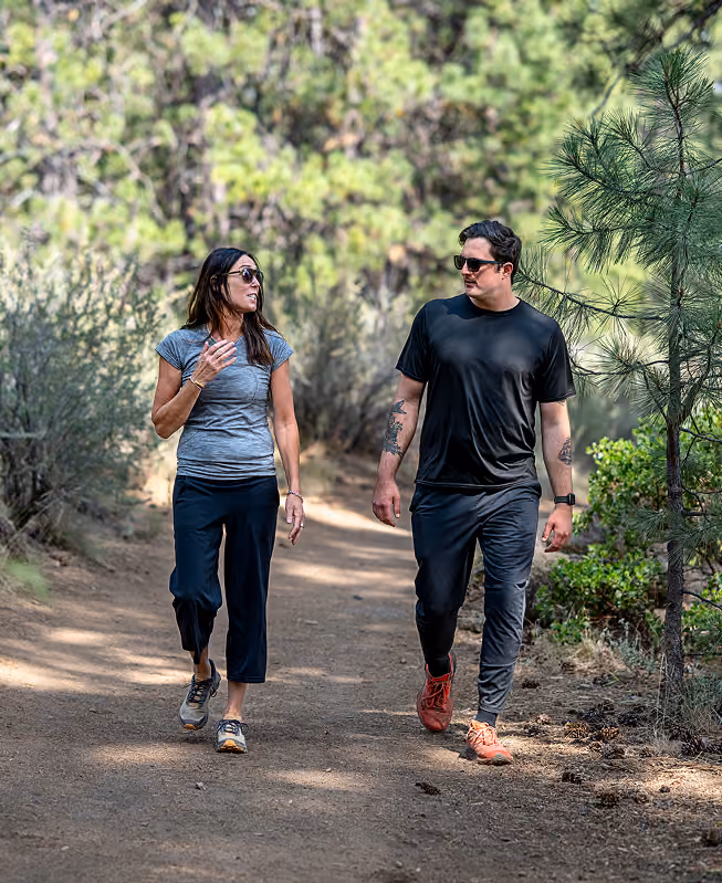A man and a woman wearing sunglasses walking and talking on a wooded trail surrounded by trees and bushes.