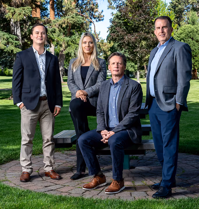Group of four professionally dressed adults posing outdoors on a circular stone patio with green grass and trees in the background; two men stand on either side, a man sits on a picnic bench, and a woman rests one foot on the bench.