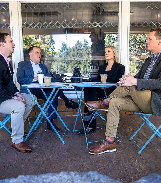 Four business professionals, two men on the left and a woman and man on the right, sitting on blue metal chairs around two blue metal tables outside a building, each with iced coffee cups, engaged in conversation.
