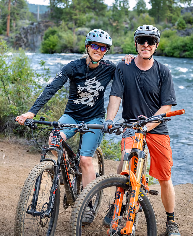 A smiling couple wearing helmets and sunglasses posing with their mountain bikes on a dirt trail by a river surrounded by green trees.
