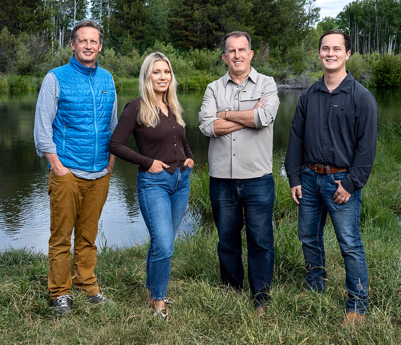 Four adults standing outdoors on grass near a calm pond with trees in the background. From left to right: a man in a blue vest and brown pants, a blonde woman in a black top and blue jeans, a man in a light gray shirt and dark jeans with arms crossed, and a man in a black shirt and blue jeans with a brown belt. All are smiling and facing forward.