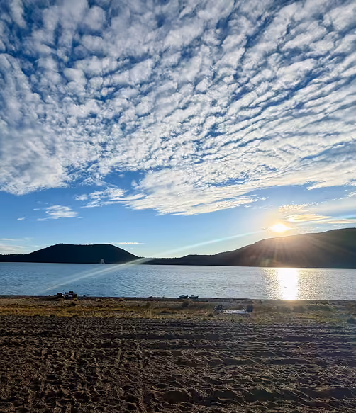 Sunset over a calm lake with hills in the background and a sandy shore in the foreground, under a sky filled with scattered clouds.
