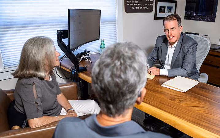 A man in a gray suit sitting at a wooden desk talking to an older woman and man seated across from him in an office setting with a computer monitor on the desk.
