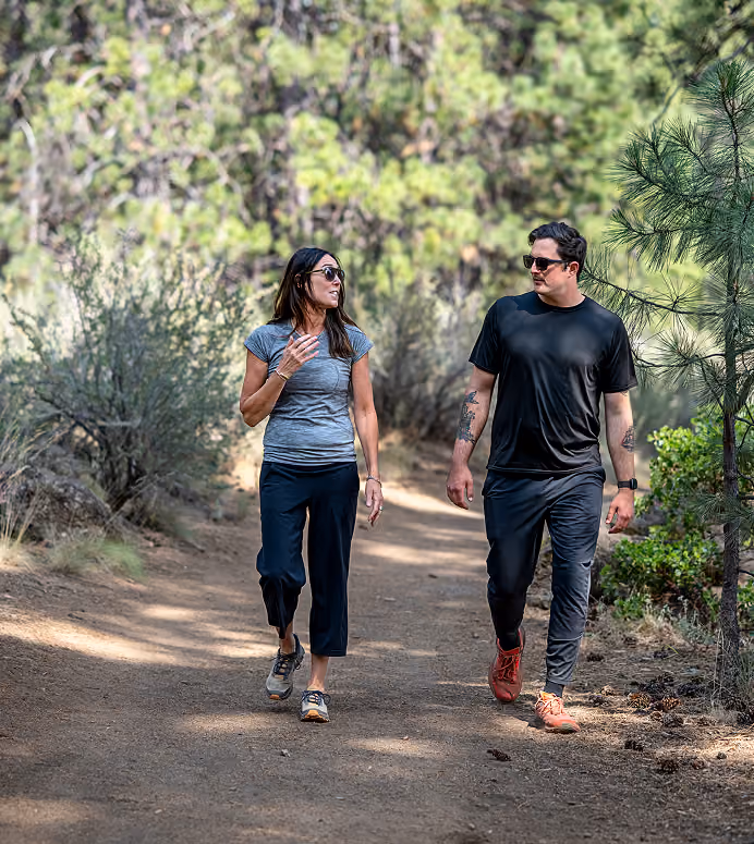 A man and a woman wearing sunglasses walking and talking on a wooded trail surrounded by trees and bushes.
