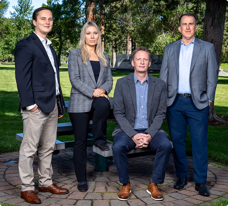 Group of four professionally dressed adults posing outdoors on a circular stone patio with green grass and trees in the background; two men stand on either side, a man sits on a picnic bench, and a woman rests one foot on the bench.