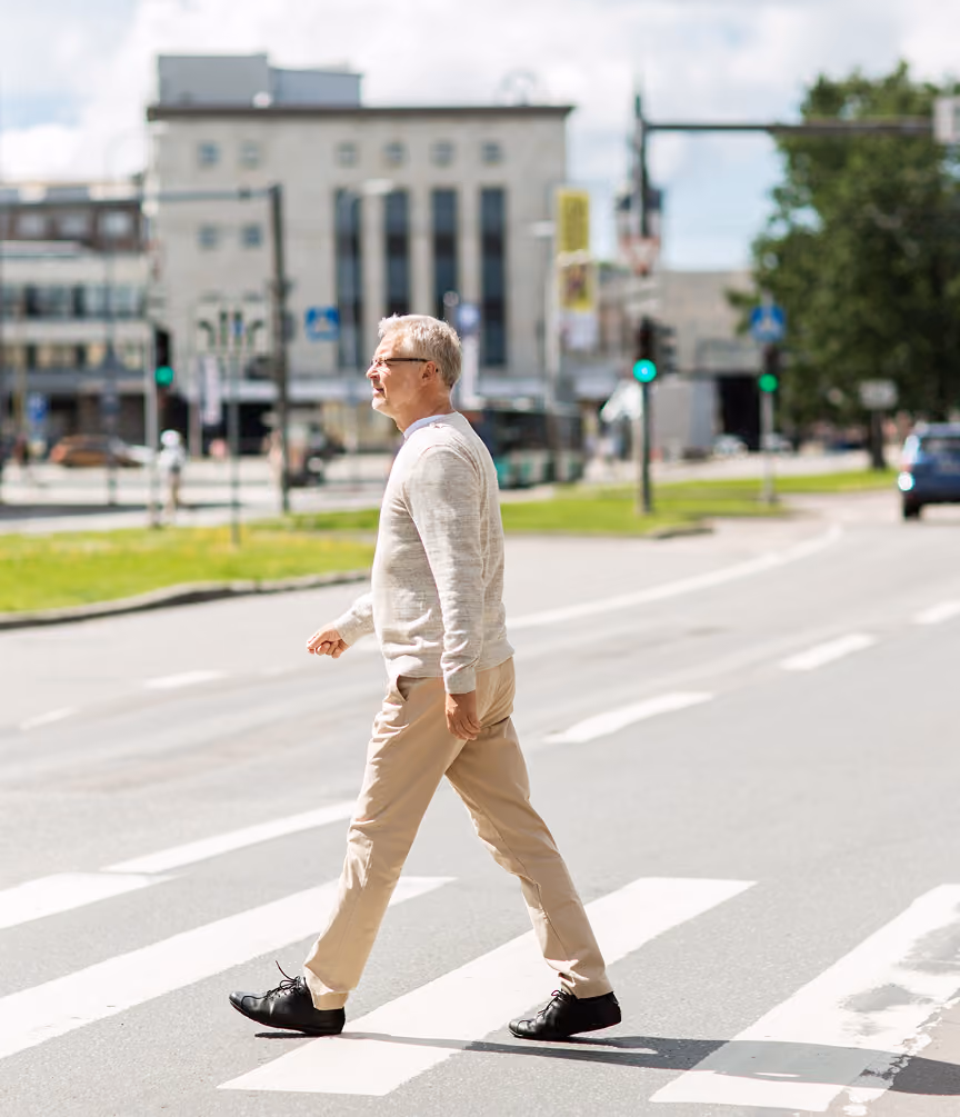 Middle-aged man with gray hair wearing a light sweater and beige pants walking across a city street pedestrian crosswalk on a sunny day.