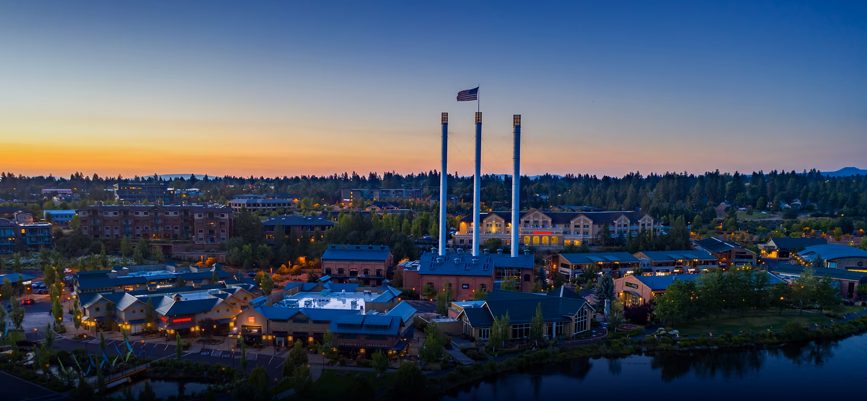 Aerial view of a Bend, Oregon at sunset with three tall smokestacks and an American flag flying atop the middle one, surrounded by buildings, trees, and a body of water reflecting the scene.