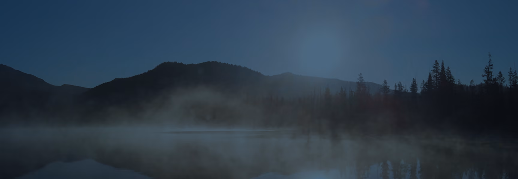 Calm lake at dusk with mist rising above the water, surrounded by dark silhouettes of trees and mountains under a deep blue sky.