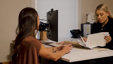 Two women at an office desk engaged in a discussion, one woman seated with documents while the other listens with folded hands.