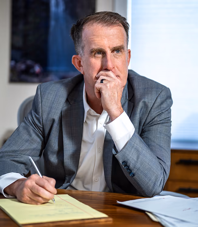 Man in a gray suit sitting at a desk, holding a pen and writing on a yellow notepad, looking thoughtfully into the distance with his hand partially covering his mouth.