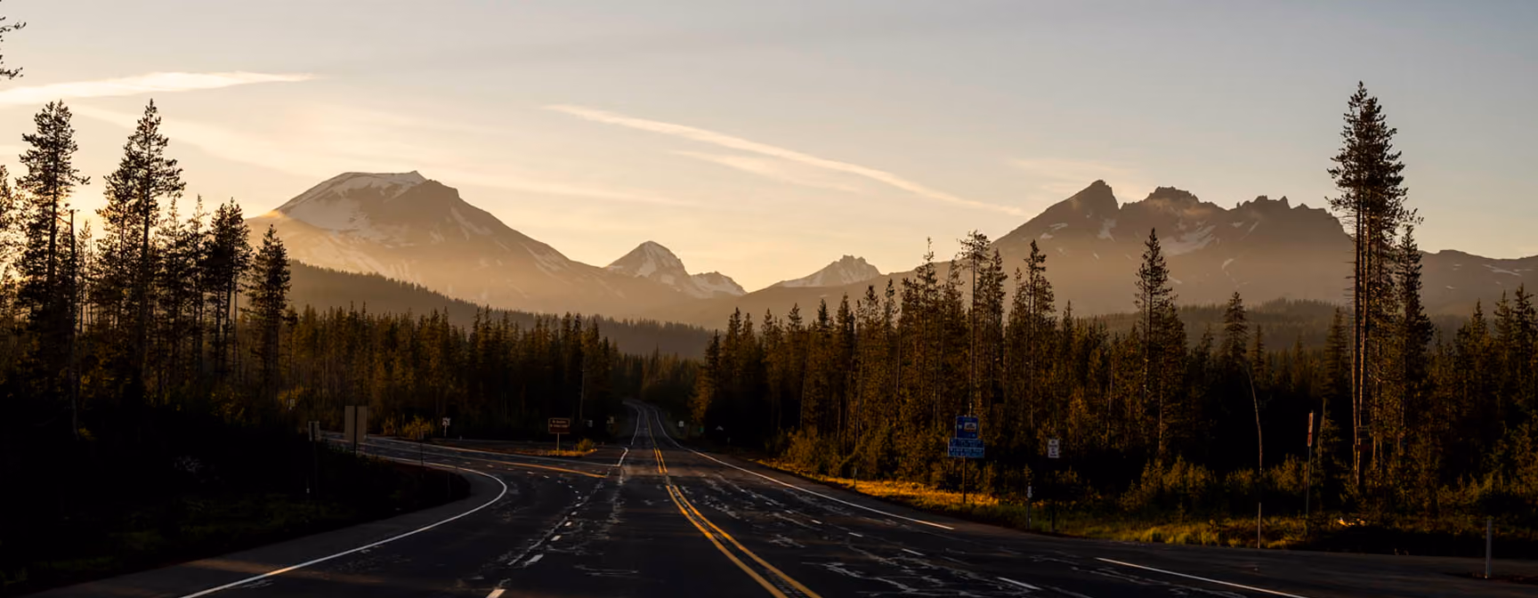 Empty paved road with yellow center lines stretches towards distant snow-capped mountains under a cloudy blue sky, flanked by tall evergreen trees.