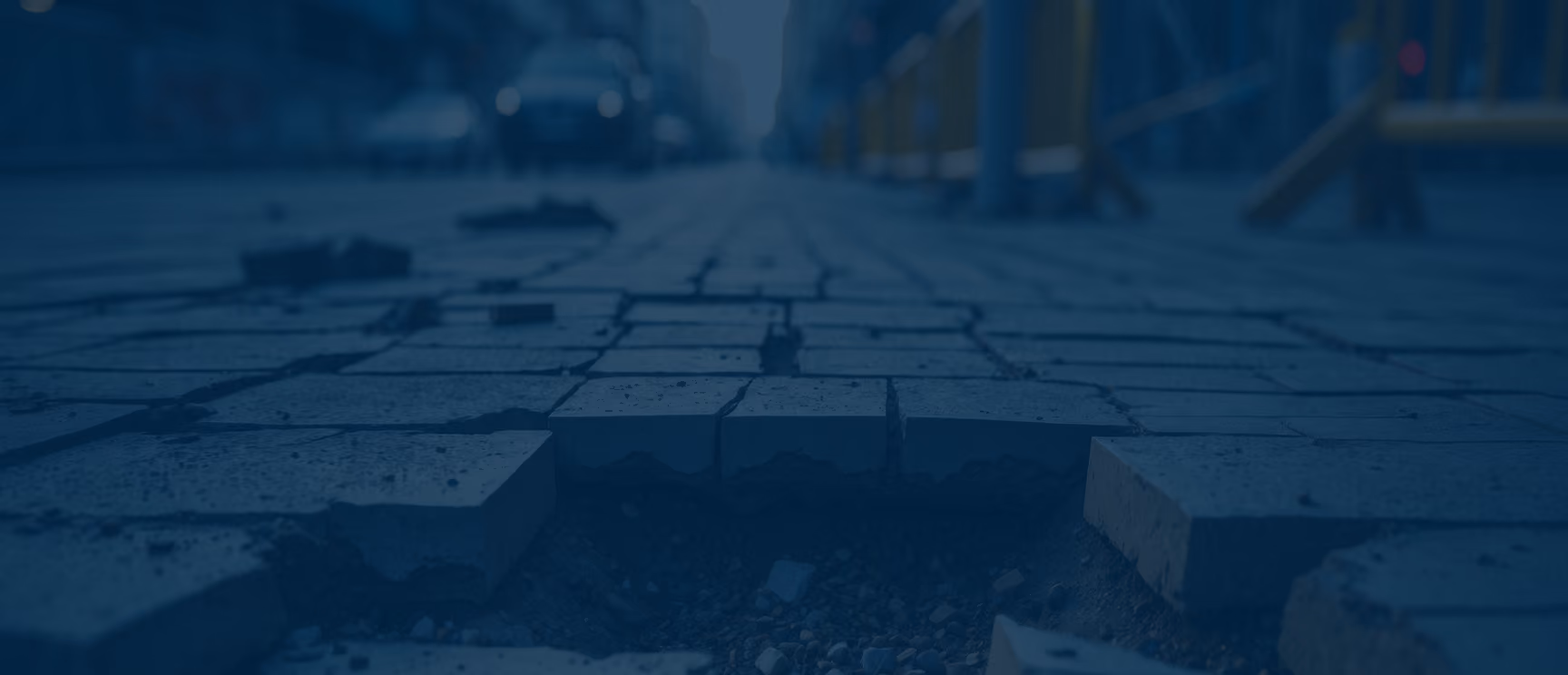 Close-up of uneven, broken bricks on a paved sidewalk with a city street blurry in the background.