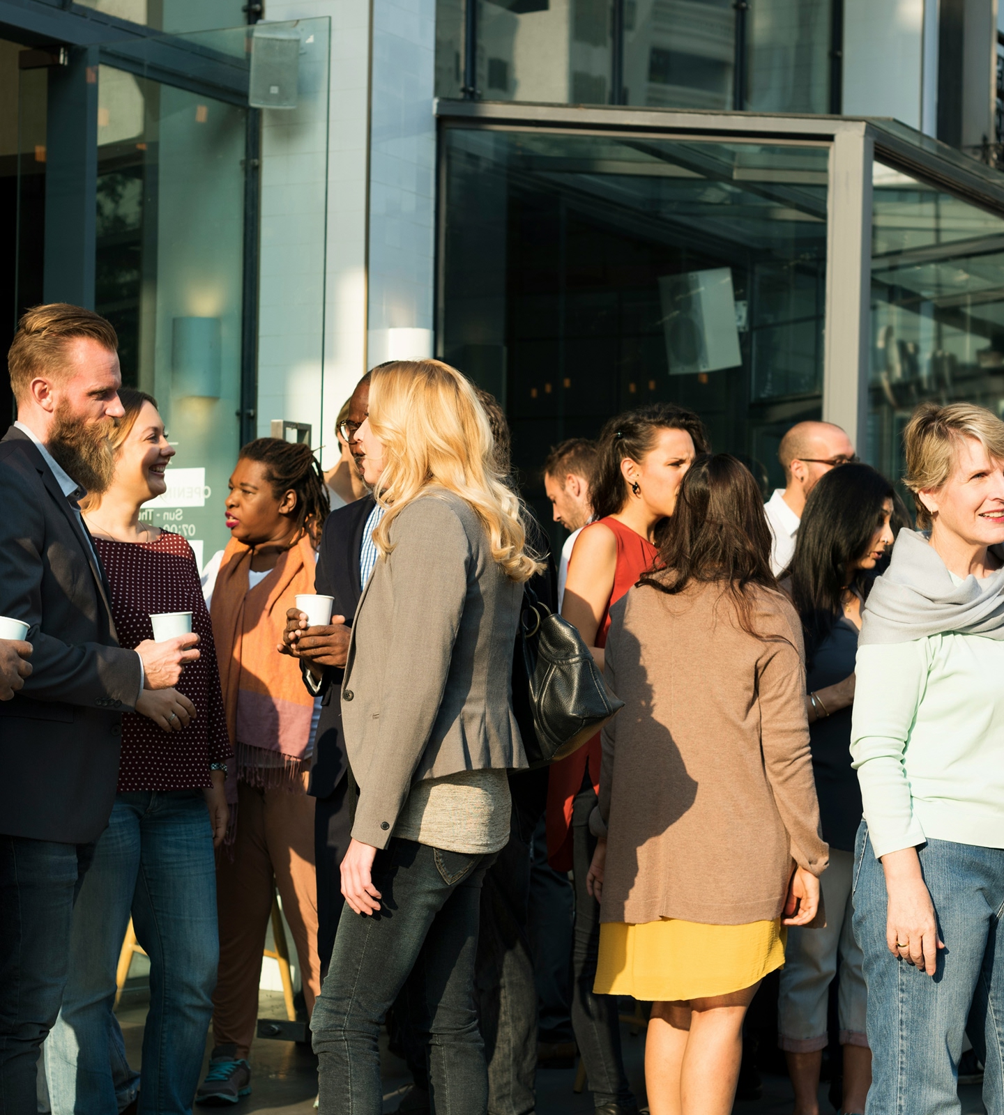 Colleagues talking in small groups with coffee outside a glass-fronted office building