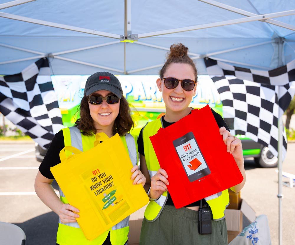 Two PRR staff in safety vests hold yellow and red tote bags printed with a 911 texting campaign message at an outdoor event.