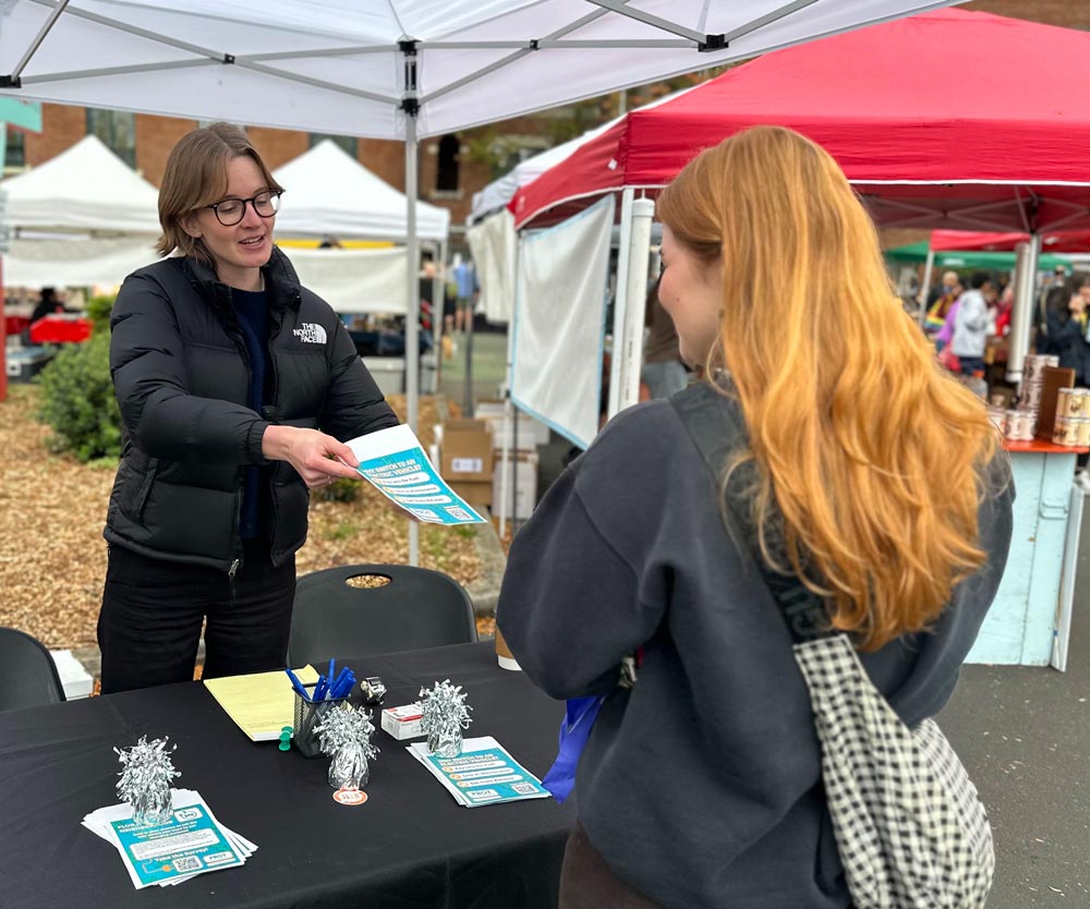 A PRR staff member hands a project flyer to a community member at an outreach table set up at an outdoor market.